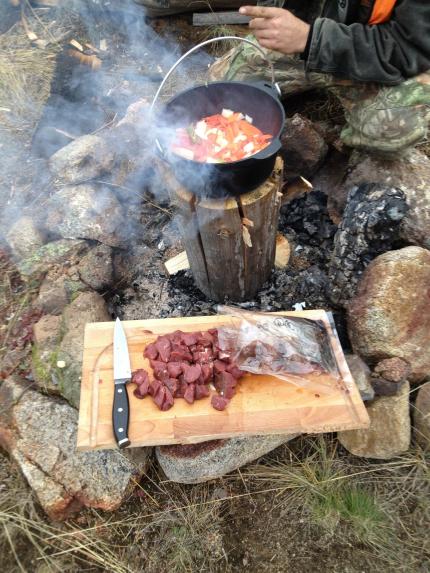 Campfire with freshly harvested game meat on cutting board, dutch oven with vegetables nearby