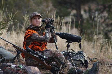 Female hunter wearing cammo and hunter orange looking through binoculars and her rifle. sitting next to a spotting scope and 